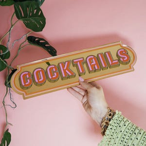 May include: A rectangular sign with the word "COCKTAILS" in orange and pink 3D lettering. The sign has a gold border and is held against a pink background. A green plant is visible in the upper left corner.