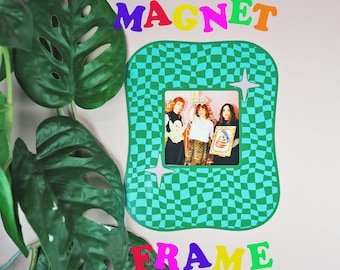 May include: A green checkered magnet frame with the word "MAGNET" in colorful letters above and "FRAME" below. The frame holds a photo of three people. Two light purple star shapes are on the frame. A green plant with large leaves is in the foreground.