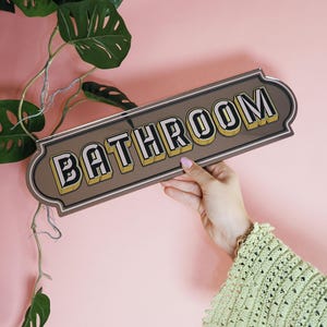 May include: A rectangular bathroom sign with the word "BATHROOM" in bold, 3D-style lettering. The sign has a brown background with a silver border and yellow accents. A hand holds the sign against a pink background.