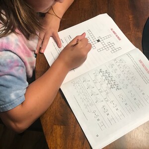 May include: A child is working on a workbook at a wooden table. The workbook pages show a crossword puzzle with the title "ALL ABOUT ALPACAS" and subtraction problems. A pencil is being used to write in the workbook.