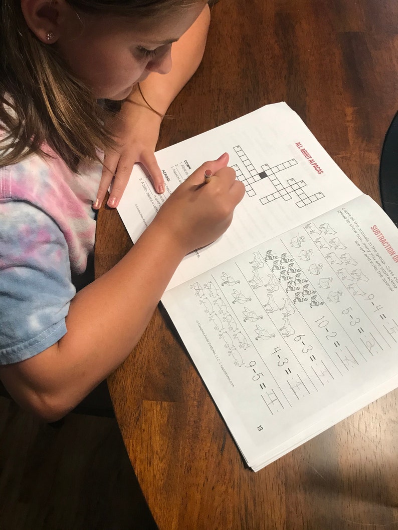 May include: A child is working on a workbook at a wooden table. The workbook pages show a crossword puzzle with the title "ALL ABOUT ALPACAS" and subtraction problems. A pencil is being used to write in the workbook.