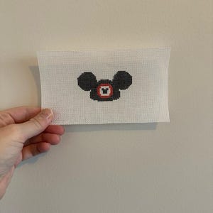 May include: A needlepoint canvas featuring a black and red Mickey Mouse design. The design is a pixelated image of Mickey's head and face, with black ears and a red and black face. The canvas is white and rectangular.