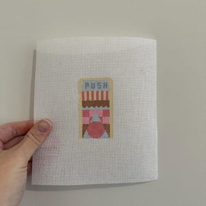 May include: A cross-stitch design on white canvas. The design features a door with the word "PUSH" above it. The door has red and pink stripes, pink bows, and a pink circle. The canvas is held by a hand.