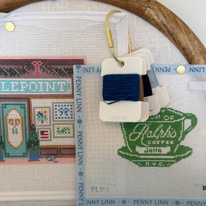 May include: A white needlepoint canvas featuring a storefront illustration and a green coffee cup graphic with the text Ralph's Coffee Java N.Y.C. A brass carabiner holds several white plastic thread bobbins wrapped in blue and grey embroidery floss.