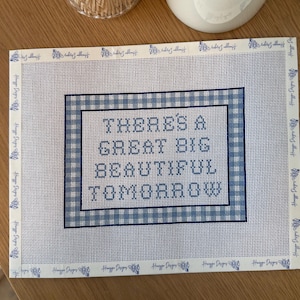 May include: A rectangular needlepoint canvas featuring a blue and white gingham border and the text "THERE'S A GREAT BIG BEAUTIFUL TOMORROW" in blue block letters. The edges of the canvas are taped with white binding tape printed with blue bees.
