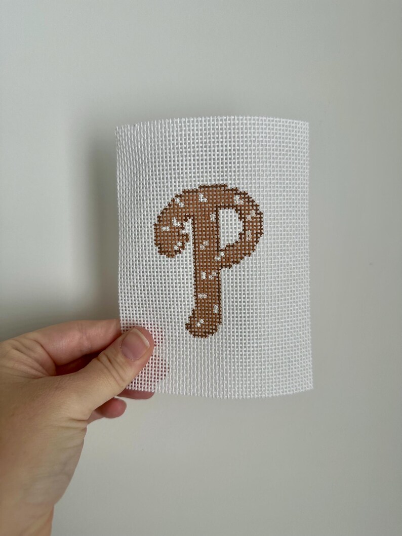 May include: A white canvas with a brown stitched letter "P" design. The letter is filled with a lighter brown and white speckles. The canvas is held by a hand against a white background.