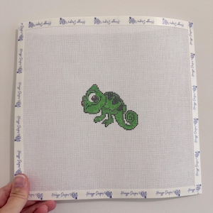 May include: A square needlepoint canvas featuring a green chameleon design centered on a white grid. The edges of the canvas are bound with white tape printed with the text Huggy Designs and a bee icon repeated along the entire border.