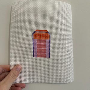 May include: A hand-stitched needlepoint canvas featuring a colorful design. The design includes a rectangular shape with the word "PUSH" in orange, pink, and purple hues. The canvas is held against a neutral background.
