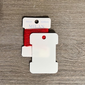 May include: Two white cardboard bobbins for embroidery thread. One bobbin is wrapped with bright red thread and features the printed text TILLI TOMAS MILAN. The second bobbin is blank and positioned partially in front of the first one.