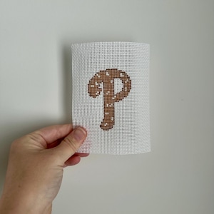 May include: A white canvas with a brown stitched letter "P". The letter is in a serif font and has small white stitches throughout. The canvas is held by a hand against a white background.