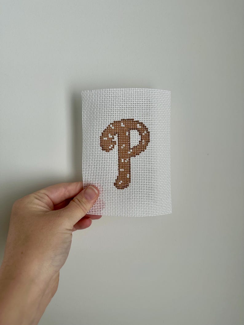May include: A white canvas with a brown stitched letter "P". The letter is in a serif font and has small white stitches throughout. The canvas is held by a hand against a white background.