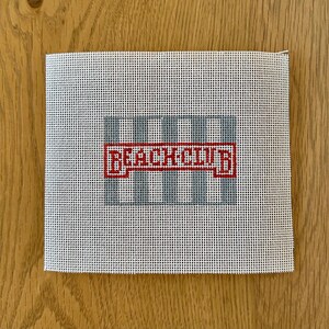 May include: A square needlepoint canvas with a grid design in light blue and the words "BEACH CLUB" in red. The canvas is white with a grid pattern, and the text is in a bold, blocky font.