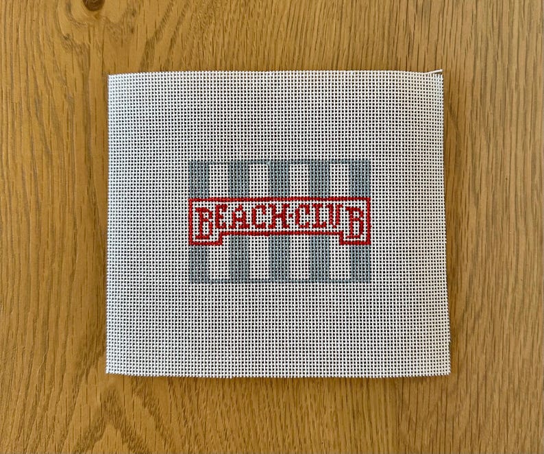 May include: A square needlepoint canvas with a grid design in light blue and the words "BEACH CLUB" in red. The canvas is white with a grid pattern, and the text is in a bold, blocky font.