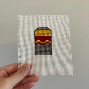 May include: A hand-stitched needlepoint design featuring a stylized image of a container. The container is primarily gray with a yellow top and a red wavy line in the middle. The needlepoint is on a white canvas.