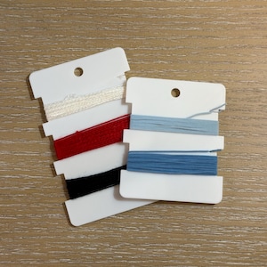 May include: Two white plastic bobbins wrapped with embroidery floss. One bobbin holds white, red, and black thread. The second bobbin, positioned slightly to the right, holds two shades of light and medium blue thread. Each bobbin has a hole at the top.