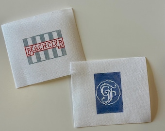 May include: Two white needlepoint canvases. One has a red and gray striped design with the words "BEACH CLUB" in red. The other has a blue square with a white monogram. Both are likely for crafting.