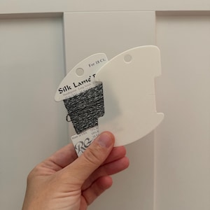 May include: A spool of silver metallic Silk Lame' thread wrapped around a white plastic card. The label reads Silk Lame' for 18 Ct. Needlepoint Long Stitch, Made in USA, 72% Silk, 10% Metallic, 10 Yards. The thread has a shimmering, textured appearance.