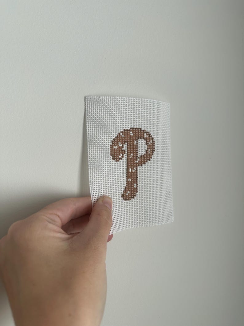 May include: A canvas with a brown letter "P" in a cross-stitch design. The letter is filled with a pattern of small white dots. The canvas is held against a white wall.