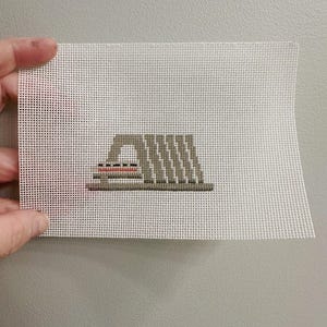 May include: A hand-stitched needlepoint canvas featuring a geometric design in shades of gray, with accents of red and black. The canvas is held against a neutral gray background, showcasing the detailed cross-stitch work.
