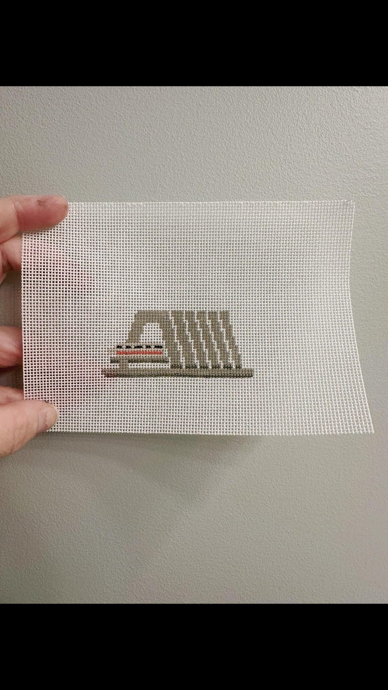 May include: A hand-stitched needlepoint canvas featuring a geometric design in shades of gray, with accents of red and black. The canvas is held against a neutral gray background, showcasing the detailed cross-stitch work.