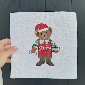 May include: Needlepoint canvas featuring a brown bear wearing a red Santa hat, green and white striped shirt, and a red apron with the name "Ralphie". The bear is holding a cup and a pair of scissors. The canvas is white.