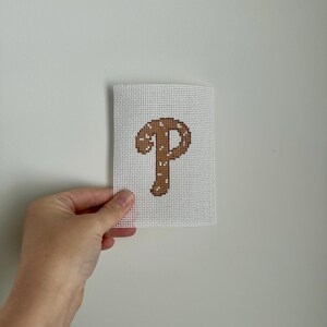 May include: A white canvas with a brown letter "P" cross-stitched in the center. The letter is decorated with small white dots. The canvas is held against a white wall.