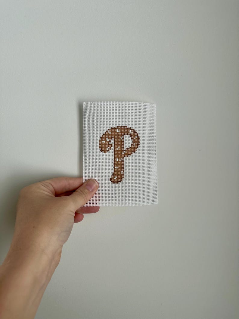 May include: A white canvas with a brown letter "P" cross-stitched in the center. The letter is decorated with small white dots. The canvas is held against a white wall.