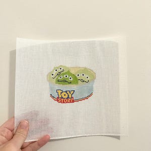 May include: Cross-stitch of the Toy Story aliens in a blue and white container. The aliens are green with three eyes. The Toy Story logo is below the aliens in yellow, red, and blue.