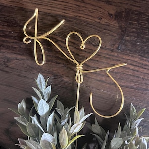 May include: Gold wire cake topper featuring the letters "K" and "J" with a heart-shaped design. The topper is placed above a bed of green leaves, creating a decorative accent for a wedding or celebration cake.