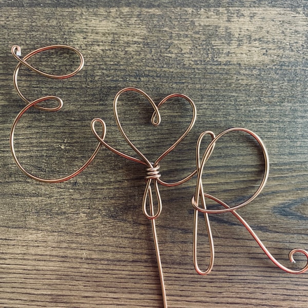 May include: A copper-colored wire sculpture featuring the cursive letter E, a central heart shape, and the cursive letter R. The wire is bent into continuous loops and curves, forming a decorative topper with a single straight wire stem extending downward.