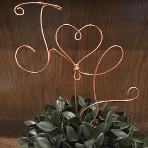 May include: A copper wire sculpture of the letters "J" and "S" intertwined with a heart shape. The sculpture is sitting on a bed of green foliage.