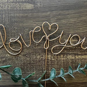 May include: Gold wire sculpture of the words "Love You" with a heart in the middle. The sculpture is on a wooden surface with green leaves.