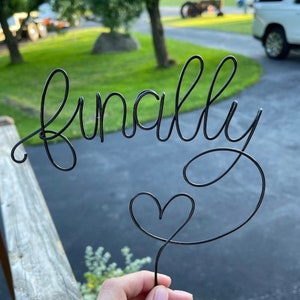 May include: A black wire cake topper featuring the word finally in a continuous cursive script, with a small heart shape integrated into the tail of the letter y. The wire is thin and dark, forming a single connected piece of decorative metal art.