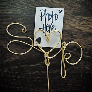 May include: Gold wire heart shaped photo holder with the words "Photo Here" written on a white card.