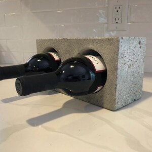 May include: A gray concrete wine rack holding two bottles of red wine. The bottles are dark with black bottle tops and white labels. The rack has two circular openings to hold the wine bottles.