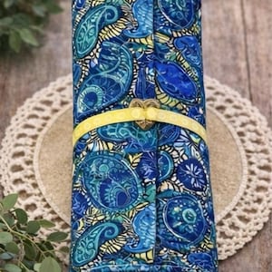 May include: A rolled-up, patterned fabric item with a blue paisley design. A yellow ribbon with a heart-shaped clasp secures the item. The fabric is a mix of blues and greens.