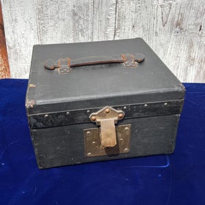 May include: A black rectangular storage box with a dark leather handle on the top lid. The box features a brass-colored metal latch and lock plate on the front center, secured with small rivets along the edges of the lid and the main body.