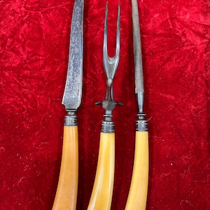 May include: A set of three vintage silver-plated serving utensils with bone handles. The set includes a carving knife, a fork, and a carving steel. The utensils are displayed on a red velvet background.