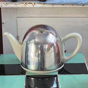 May include: A silver-colored teapot with a hammered texture, a white spout, and a white handle. The teapot sits on a black, white, and turquoise tiled surface. The teapot has a rounded body and a flat base.