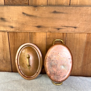 May include: An oval copper cooking pot with two brass handles and a matching oval lid featuring a central brass handle. The copper surface displays a natural patina with visible variations in color and texture across the metal exterior.