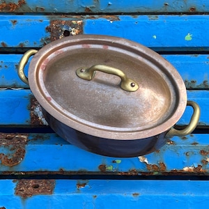 May include: An antique copper cooking pot with a lid. The pot has brass handles and a brass handle on the lid. The pot is oval-shaped and sits on a blue surface with rust spots.