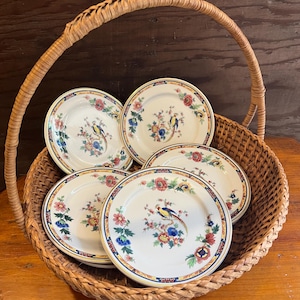 May include: A set of five white ceramic plates with a floral and bird design, displayed in a woven wicker basket. The plates feature a yellow border with a blue and red floral pattern and a colorful bird motif.