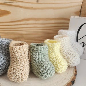 Op de afbeelding: Een verzameling handgehaakte babyslofjes in verschillende kleuren, waaronder grijs, beige, groen, geel en wit. De slofjes zijn gerangschikt op een houten schijf, met een bordje met de tekst "BE BRAVE LITTLE ONE".