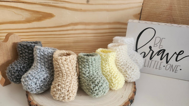 Op de afbeelding: Een verzameling handgehaakte babyslofjes in verschillende kleuren, waaronder grijs, beige, groen, geel en wit. De slofjes zijn gerangschikt op een houten schijf, met een bordje met de tekst "BE BRAVE LITTLE ONE".