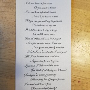 Op de afbeelding: Een witte rechthoekige kaart met een gedicht in sierlijke letters. Het gedicht drukt anticipatie en liefde uit, waarschijnlijk voor een pasgeboren baby. De kaart heeft een subtiele glans en ligt op een houten oppervlak.