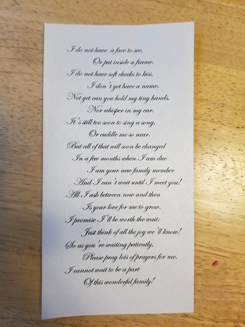Op de afbeelding: Een witte rechthoekige kaart met een gedicht in sierlijke letters. Het gedicht drukt anticipatie en liefde uit, waarschijnlijk voor een pasgeboren baby. De kaart heeft een subtiele glans en ligt op een houten oppervlak.