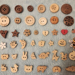 Op de afbeelding: Een verzameling houten knopen in verschillende vormen en maten. Bevat ronde knopen in bruine tinten en decoratieve knopen met hart-, ster- en diermotieven. Sommige knopen hebben de tekst "Little Angel" en "Handmade with love".