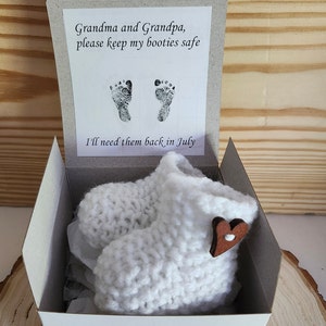 Op de afbeelding: Witte gebreide babyslofjes met een houten hartaccent, gepresenteerd in een witte doos met de tekst: "Oma en Opa, bewaar mijn slofjes alsjeblieft. Ik heb ze in juli weer nodig."