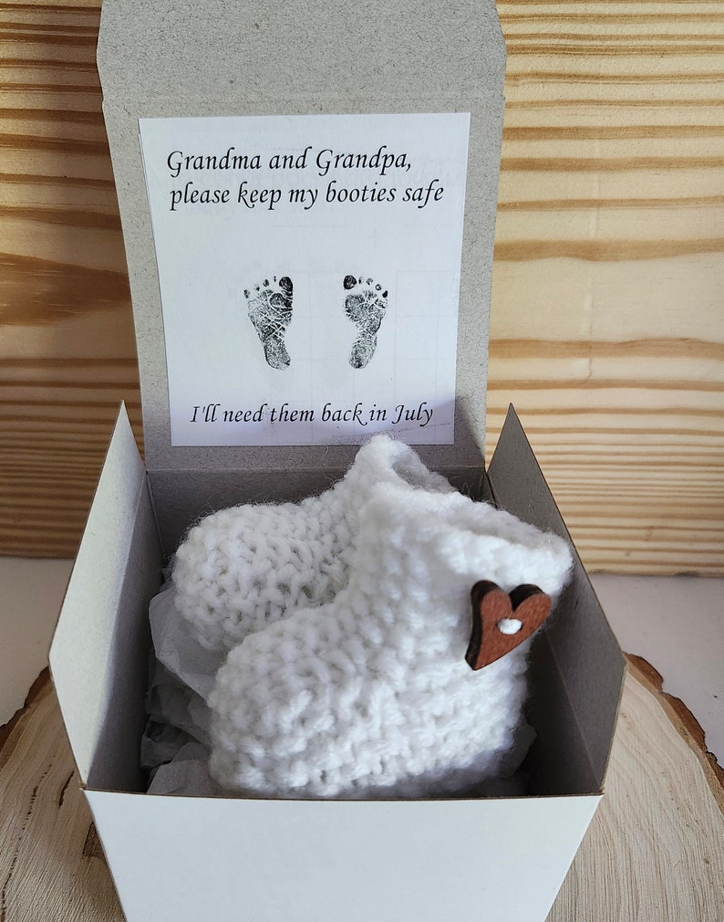 Op de afbeelding: Witte gebreide babyslofjes met een houten hartaccent, gepresenteerd in een witte doos met de tekst: "Oma en Opa, bewaar mijn slofjes alsjeblieft. Ik heb ze in juli weer nodig."