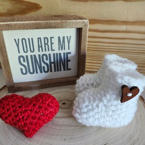 Op de afbeelding: Een witte gehaakte babyschoen met een houten hartknop, een rood gehaakt hart en een ingelijst bord met de tekst "YOU ARE MY SUNSHINE". De items worden gepresenteerd op een houten oppervlak.